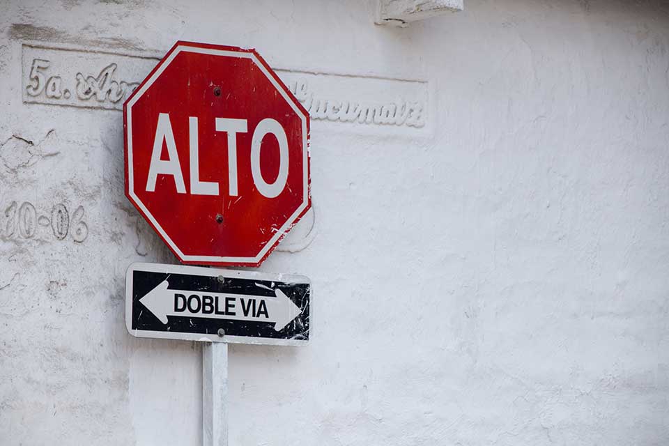 A stop sign displayed up against a white wall. Sign reads (in Spanish): Alto. Doble Via.