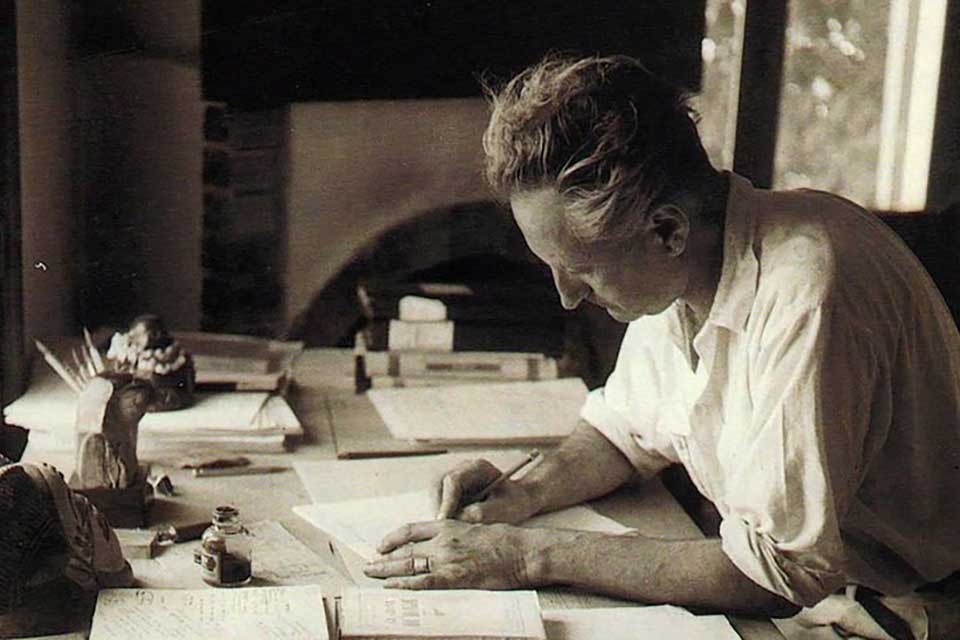 A sepia-toned photograph of Jean Giono sitting at a cluttered desk writing