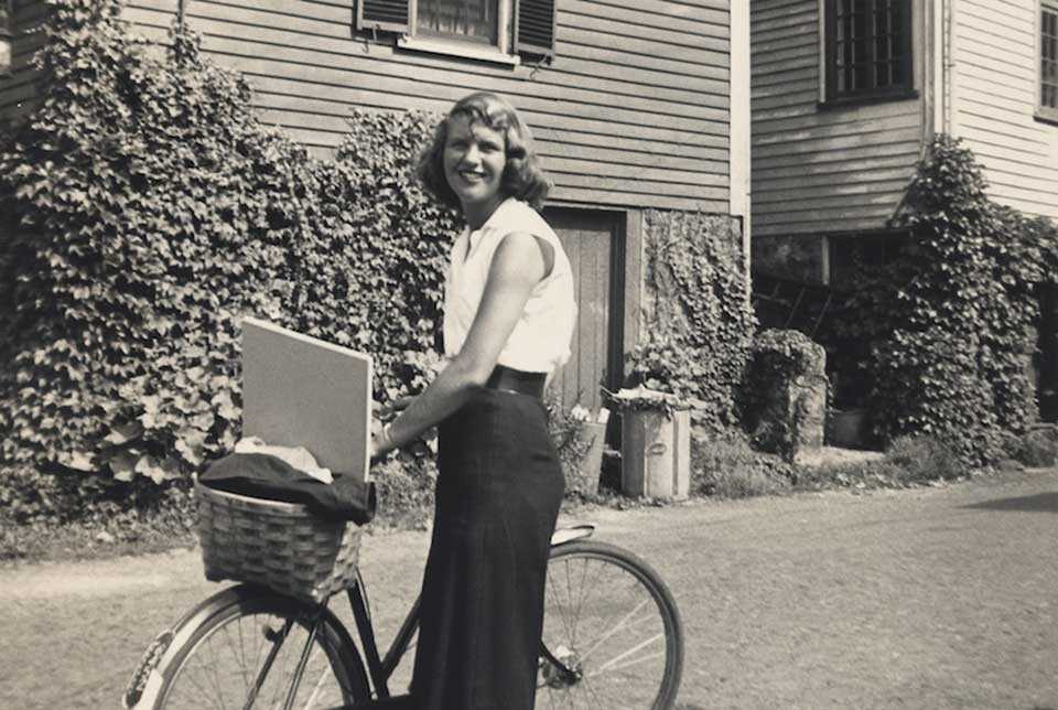 A black and white photo of Sylvia Plath with her bicycle smiling at the camera