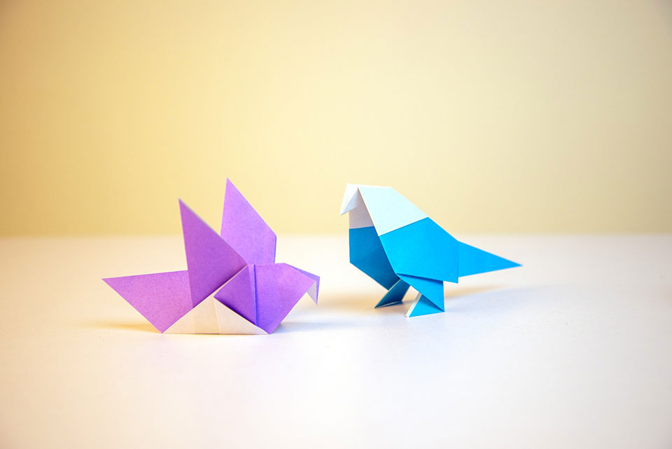 Two delicate origami pieces in the shape of birds photographs against a pale yellow background