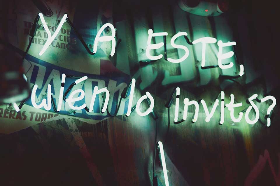 A neon sign glows in the shadows of night. The text reads 'Y A Este wién lo invito"