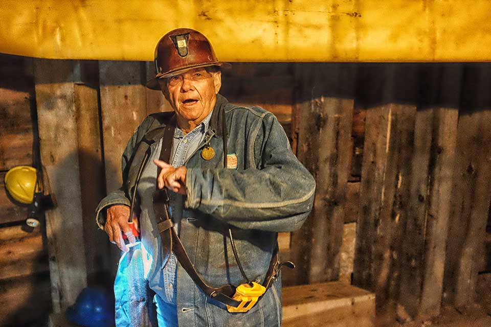 An older man in a hard hat and work coveralls appears as if he is speaking in a highly industrial location