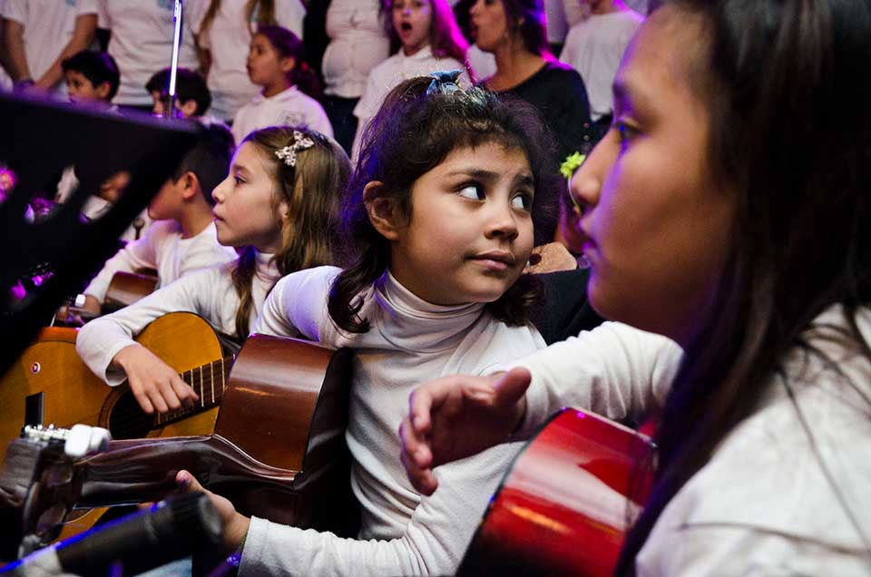 A young girl holding a guitar and surrounded by other children with instruments looks behind her