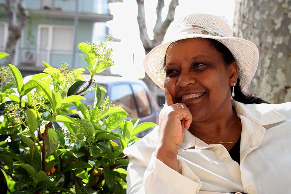 Ribka Sibhatu sits in a white suit and hat in front of a bush