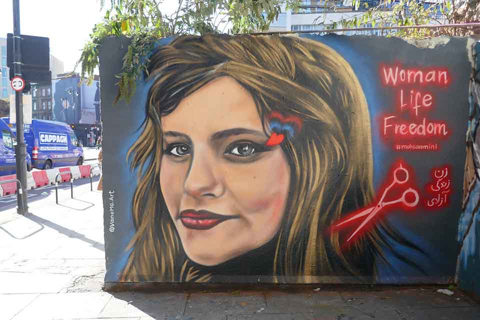 A mural of a woman's face with a pair of scissors near her head rendered in dark red. Text reads: Woman. Life. Freedom. 