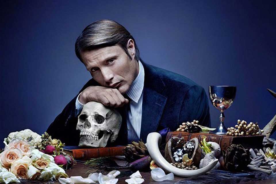 A promotional still of an actor, seated at a table, resting his chin atop his hand, which rests on a human skull