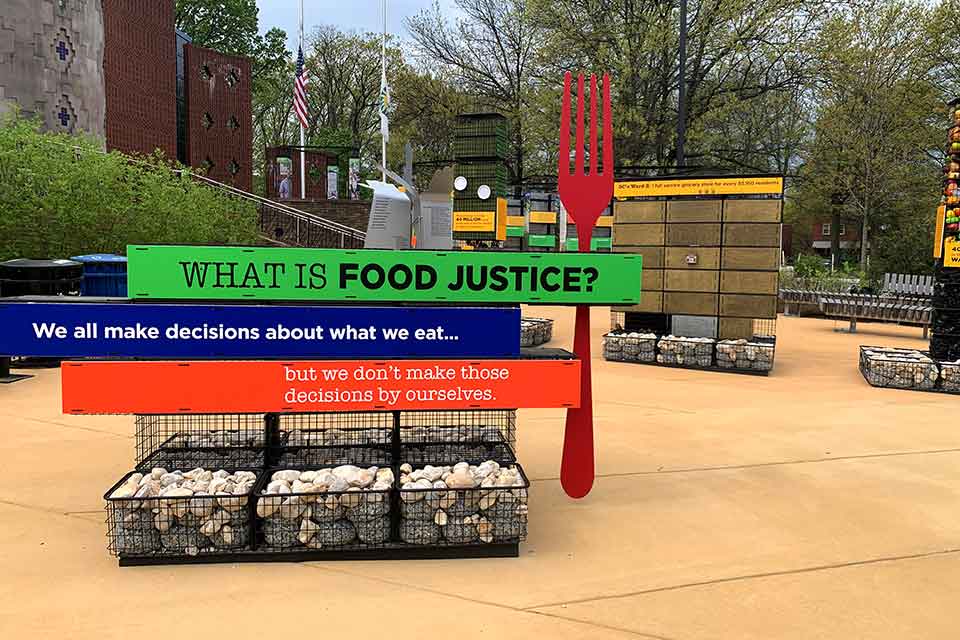 A photograph of an outdoor art installation. In the foreground, an oversized fork stands besides signs that read, "What is food justice? We all makes decisions about what we eat...but we don't make those decisions by ourselves.”