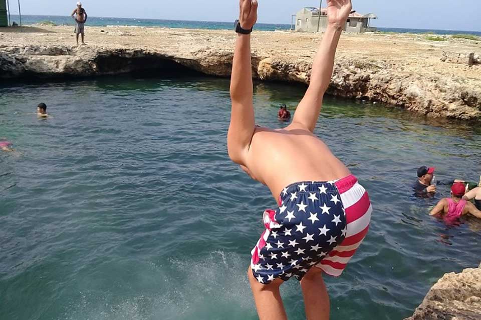 A figure clad in swimming shorts with an American flag on them faces away from the camera and is poised to jump into a natural swimming hole