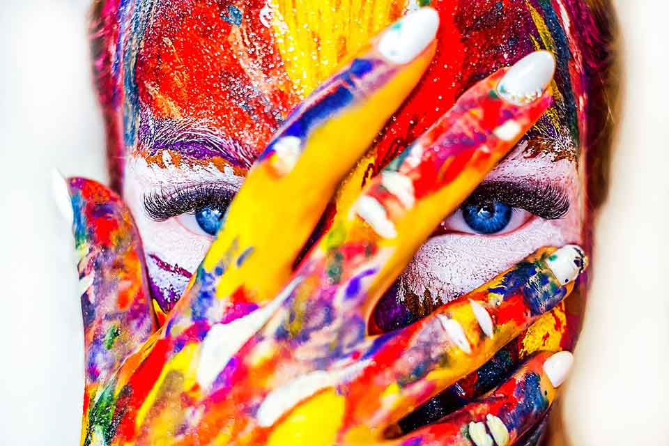 A close up on a female face streaked with a variety of different colored paints, her hand (also covered) is over her face but her fingers are split so her eyes are visible.