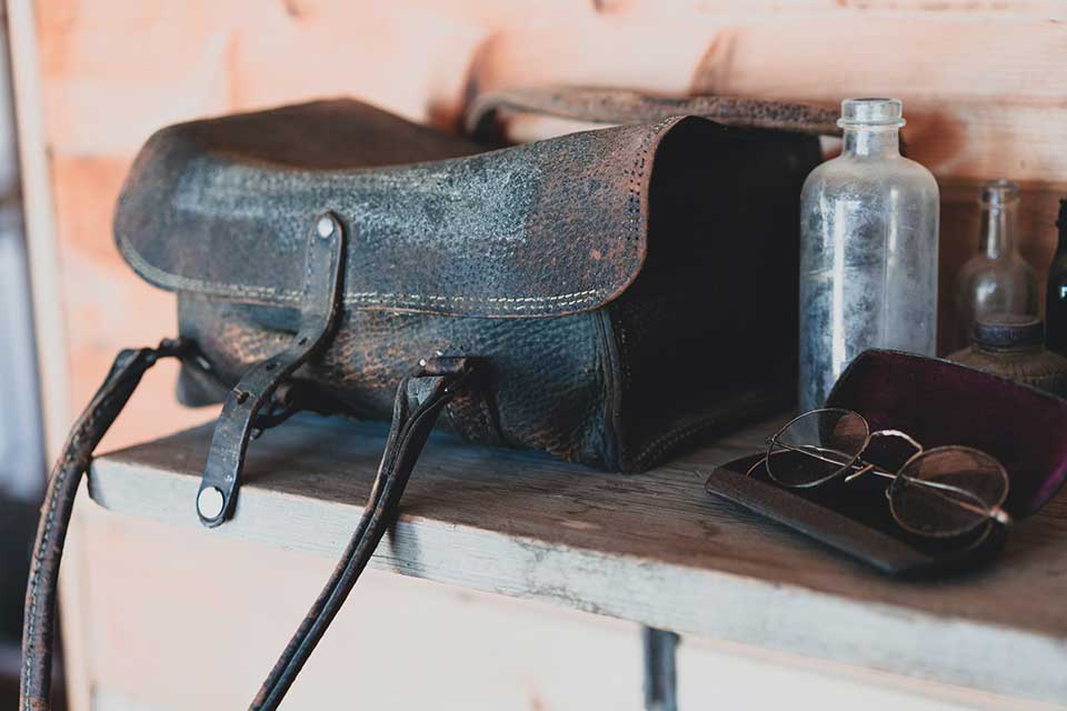 A weathered leather bag sits on a dresser with a dusty glass bottle and a pair of antique glasses sitting nearby