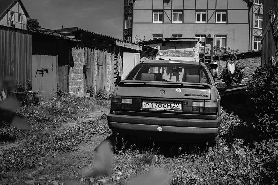 A black and white photograph of an old car parked behind a ramshackle apartment building in an overgrown alley