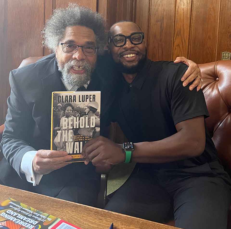 A photograph of Cornell West and Karlos Hill embracing while seated on a couch