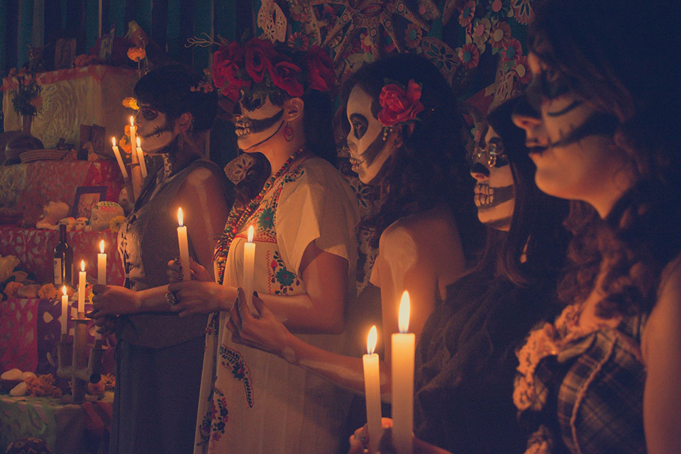 A group of people with their faces painted white like skulls hold candles before an altar