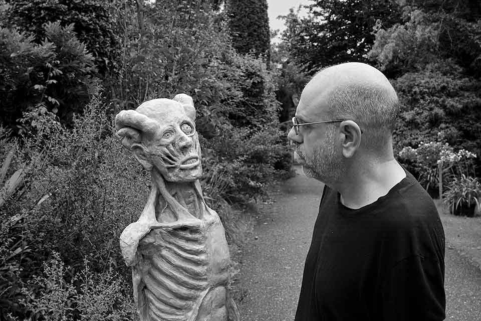 A black and white photograph of Dimitris Lyacos, who is standing on a road running down a wooded lane. He is looking at a grotesque statue that looks like a human with its skin removed.