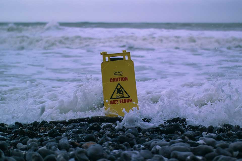 A "Caution: Wet Floor" sits on a beach and is enveloped by the tide