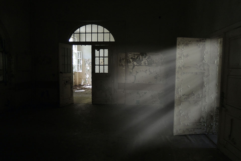 A photograph of a rundown room, mostly shrouded in shadow, with light streaking in an open door on the right