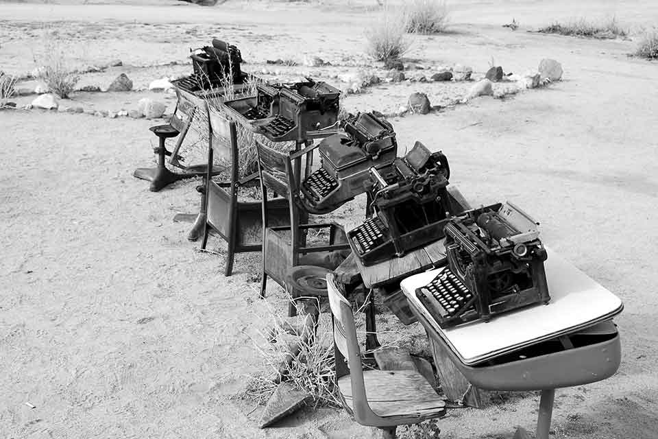 A black and white photograph of a line of typewriters on wooden desks on a beach. The desks and typewriters are covered with flotsam and sand.