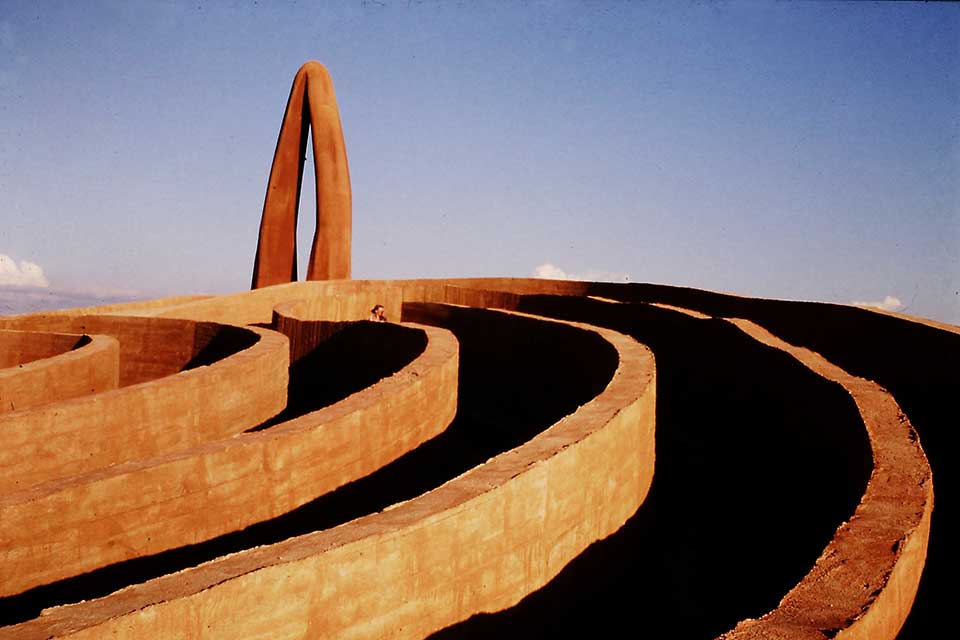 A photograph of an outdoor installation. This detail shows one edge of a series of low, concentric walls, the interiors in shadow, with a tall arc standing in the background