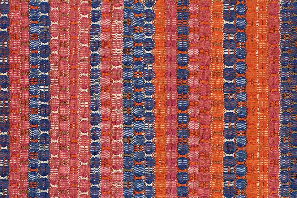 A photograph of a textile piece by Anni Albers. Colored bands (blue, yellow) travel downward against a red background