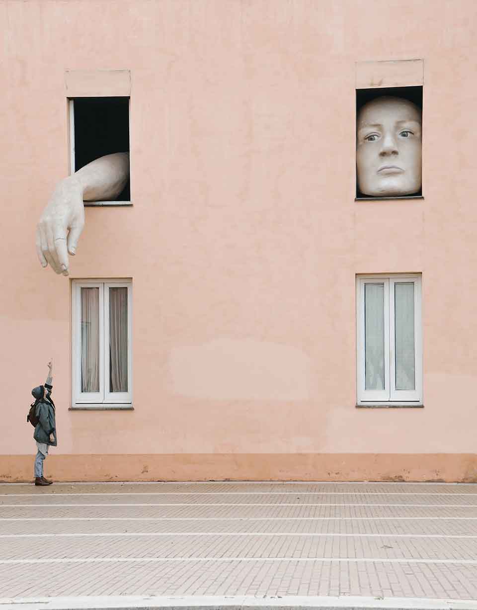 A photograph of two windows in a two story apartment building. A face fills one and an oversized hand reaches out of the second. A human figure reaches up to the hand from below