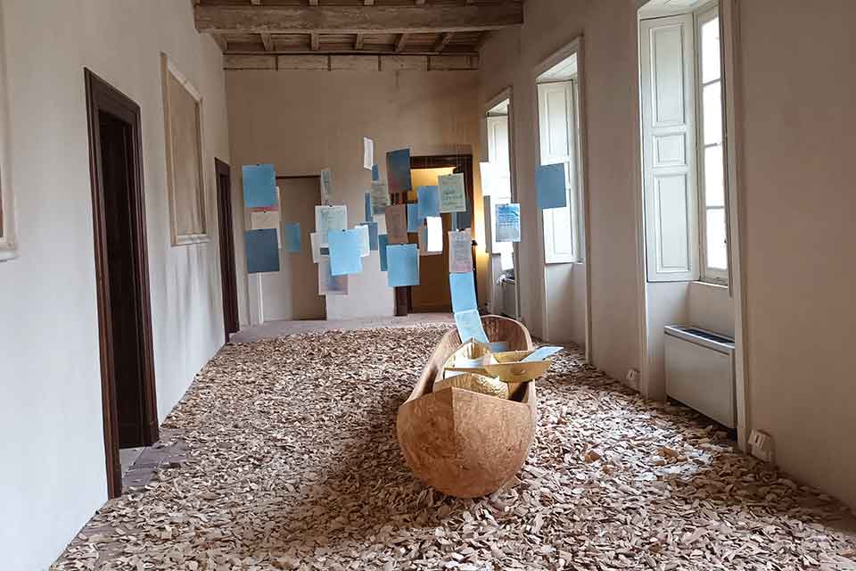 An elaborate installation. A wooden canoe sits in the middle of a room, the floor of which is covered in wood chips. Colored pieces of paper hang suspended in mid-air on string.