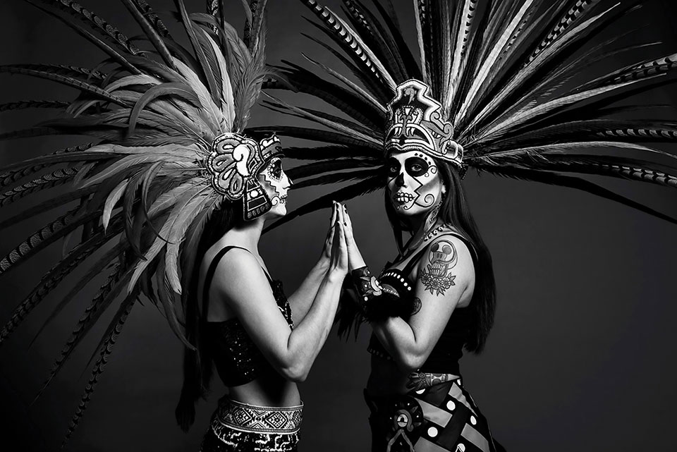 Two indigenous people, dressed in ceremonial makeup and dress, stand facing one another, palms touching