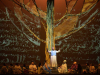 A scene from the play Omar. A man in African garb stands at the base of an immense tree, surrounded by others seated at his feet. The backdrop behind them is covered in projected Arabic text.