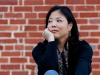 A photograph of Nicole Chung sitting in front of a red brick wall. She is looking off into the distance. 