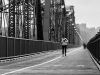 A black and white photograph of a person jogging toward the camera on a long suspension bridge