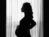 A pregnant woman standing in profile, mostly in silhouette, in front of a draped window 