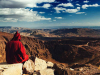 A photo of a figure, cloaked in red, sitting atop a mountain overlooking a wide vista below