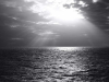 A black and white photograph of the sun peeking through the clouds above the ocean