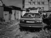 A black and white photograph of an old car parked behind a ramshackle apartment building in an overgrown alley
