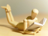 A close-up photograph of a small, wooden figure reading a tiny book.