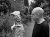 A black and white photograph of Dimitris Lyacos, who is standing on a road running down a wooded lane. He is looking at a grotesque statue that looks like a human with its skin removed.