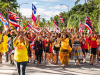A group of people, clad in brightly colored clothes, march toward the camera waving the Hawai'ian flag