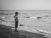 A somber black and white photograph of a boy standing on a beach looking at the ocean