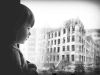 A black and white photograph of a child looking through a frosted window at a battle-damaged building across the street