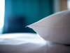 A close-up photo of a white pillow lying on a bed. The blue curtains can be seen, blurred, in the background with a hint of light from the outside streaming in.