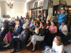 A photograph of an attentive audience, mostly seated in chairs, attending a reading in a bookstore