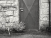 Black and white photograph of a rustic building with a padlock on the door. A scythe leans against the wall just to the left of the door