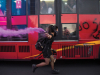 A woman masked in a black bandana runs alongside a bus covered with graffiti carrying a purple flare