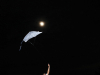 A photograph of a white kite held by an arm that stretches into the bottom of the frame with the moon hovering just above the kite