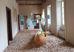 An elaborate installation. A wooden canoe sits in the middle of a room, the floor of which is covered in wood chips. Colored pieces of paper hang suspended in mid-air on string.