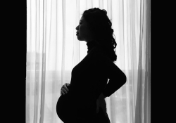 A pregnant woman standing in profile, mostly in silhouette, in front of a draped window 