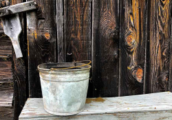 A photograph of an old galvanized steel bucket positioned in front of a wooden wall