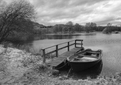A black and white photo of a small boat tied to a dock at the edge of a wooded pond