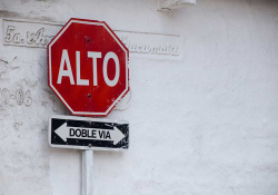 A stop sign displayed up against a white wall. Sign reads (in Spanish): Alto. Doble Via.
