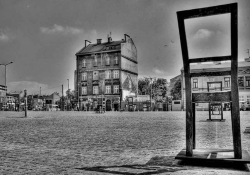 A black and white photograph of an empty town's square, with a chair that faces away from the viewer dominating the right side in the foreground