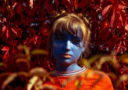 A photograph of a young woman, painted blue, centered in the frame against a oversaturated field of red leaves
